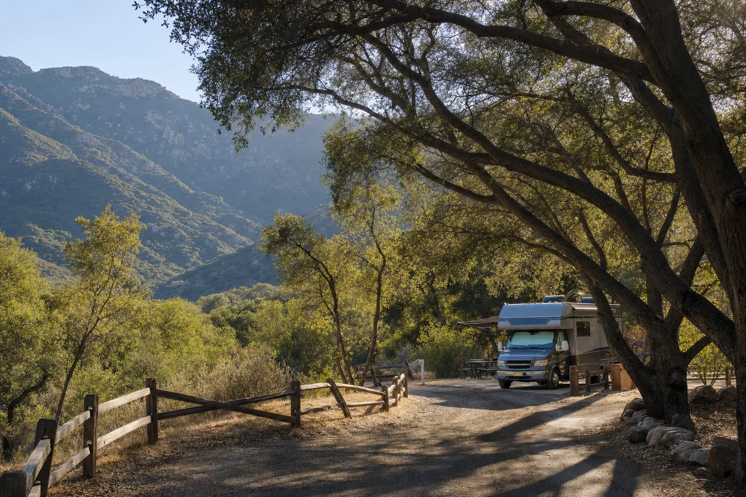 A campground lane winding beneath ancient oak trees with an RV parked in the shade and chaparral mountains behind